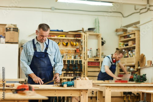 Portrait of two carpenters working with wood standing at tables in workshop, focus on senior carpenter in foreground, copy space