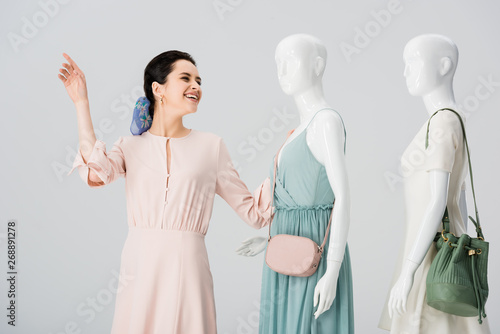 beautiful smiling girl gesturing near mannequins in dresses isolated on grey