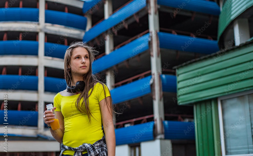 Outdoor portrait of young teenager brunette girl with long hair. Teen Girl Drinking a Bottle of fresh orange Juice on parking Background on city Outdoor portrait of young teenager brunette girl with long hair. Teen Girl Drinking a Bottle of fresh orange Juice on parking Background on city