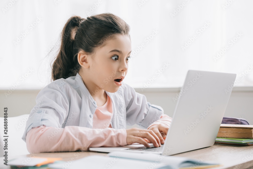 surprised child using laptop while sitting at desk and doing schoolwork at home surprised child using laptop while sitting at desk and doing schoolwork at home