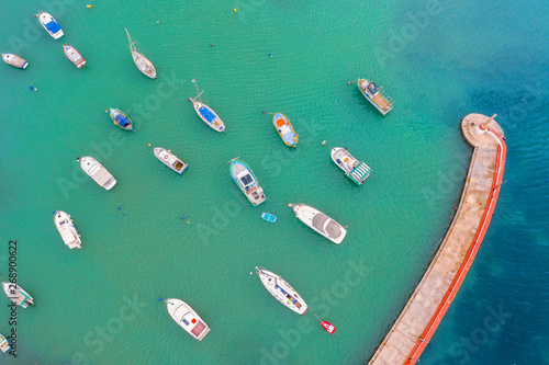 Aerial view of the bay with turquoise water and many small fishing boats with jetty and lighthouse.