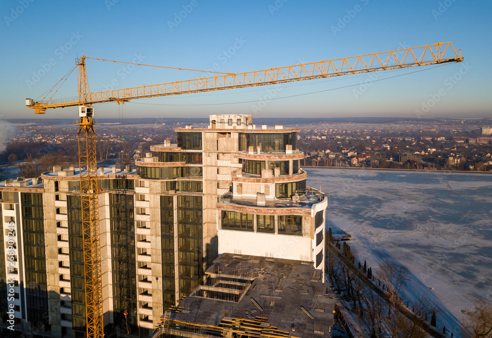 Apartment or office tall building under construction, top view. Tower crane on bright blue sky copy space background, city landscape stretching to horizon. Drone aerial photography.