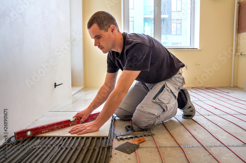 Fotografija Young worker tiler installing ceramic tiles using lever on cement floor with heating red electrical cable wire system
