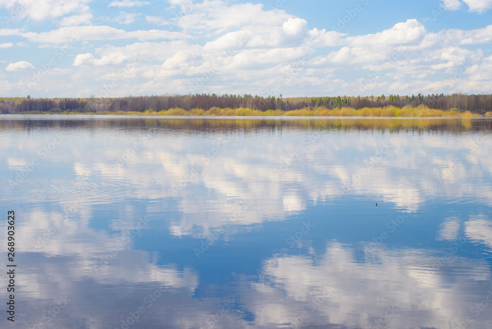 Blue sky and clouds reflected in the river. Ripples on the surface of the water. Forest on the horizon