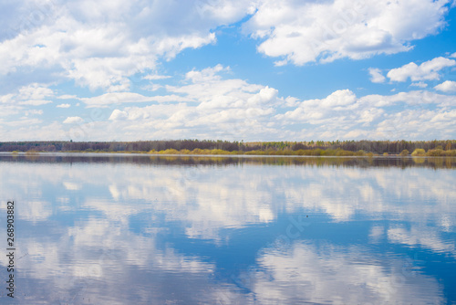 Blue sky and clouds reflected in the river. Ripples on the surface of the water. Forest on the horizon