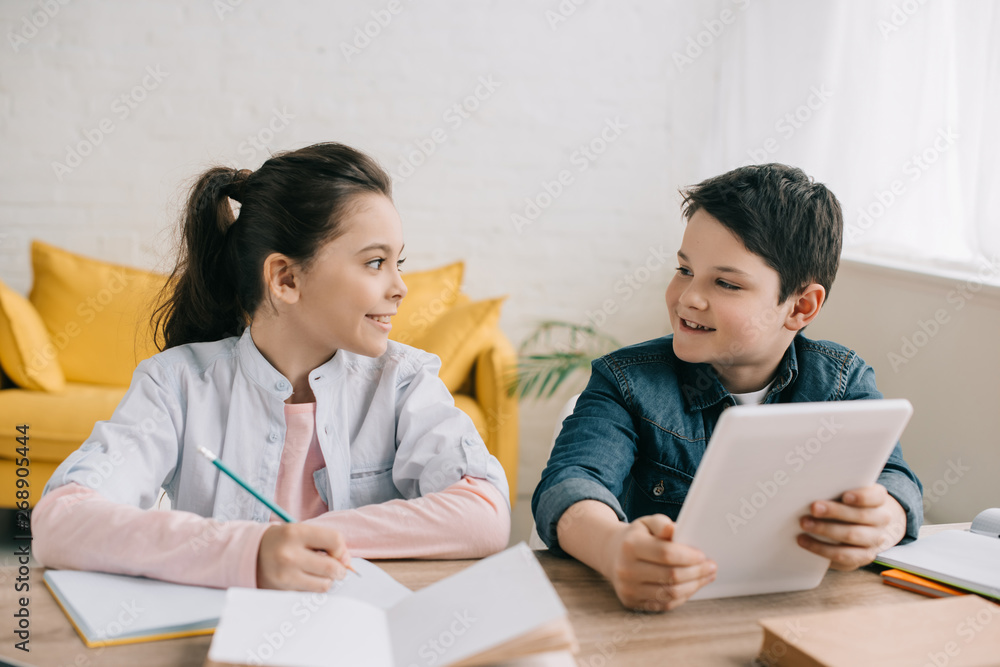 smiling boy using digital tablet while sitting at desk near sister writing in notebook smiling boy using digital tablet while sitting at desk near sister writing in notebook