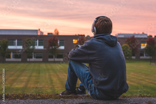 Teenager sitting alone at the top of a hill at sunset. He is listening to music through his headphones.