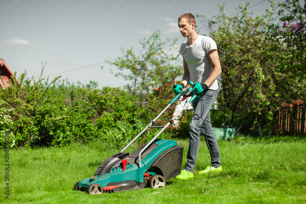 Young man mowing the lawn. Worker doing his job in backyard. Spending summer day in garden. Lawnmower standing on the background of private garden. Young man mowing the lawn. Worker doing his job in backyard. Spending summer day in garden. Lawnmower standing on the background of private garden.