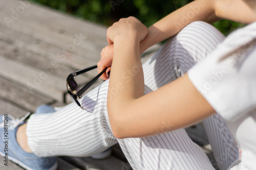 Girl hands holding sunglasses on legs with striped pants and sitting on a bench in the park on spring sunny day. Relaxation and enjoy businesswoman concept. Close up, selective focus