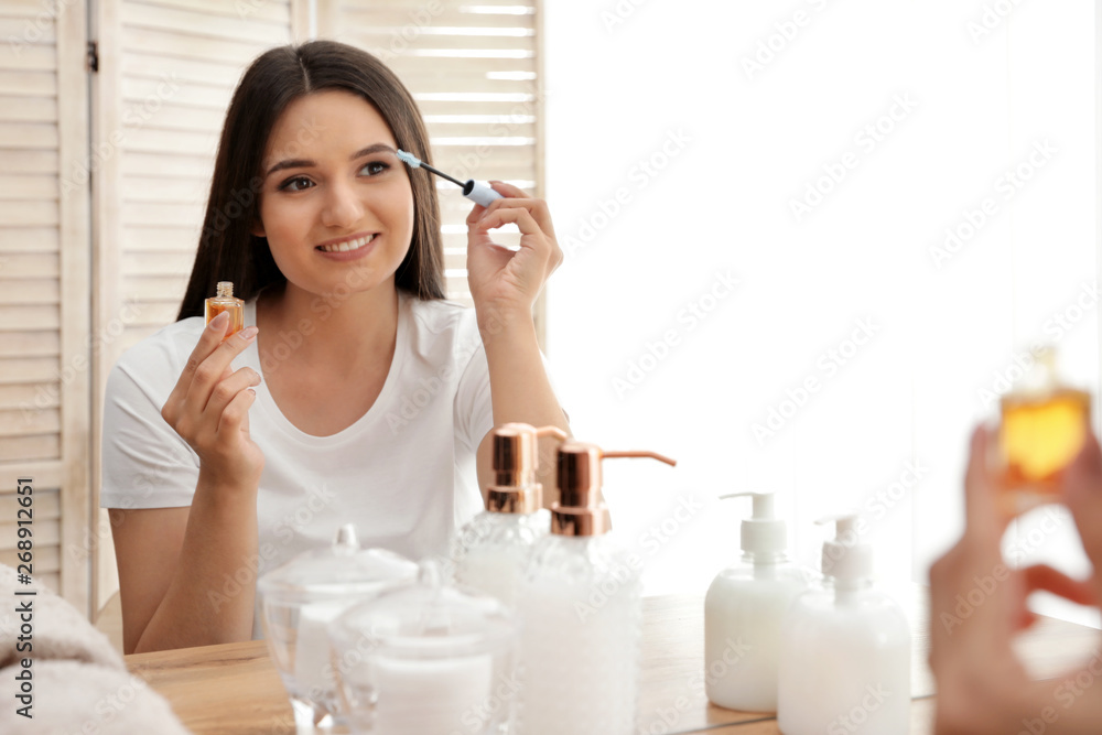 Beautiful woman applying oil onto her eyelashes near mirror indoors Beautiful woman applying oil onto her eyelashes near mirror indoors