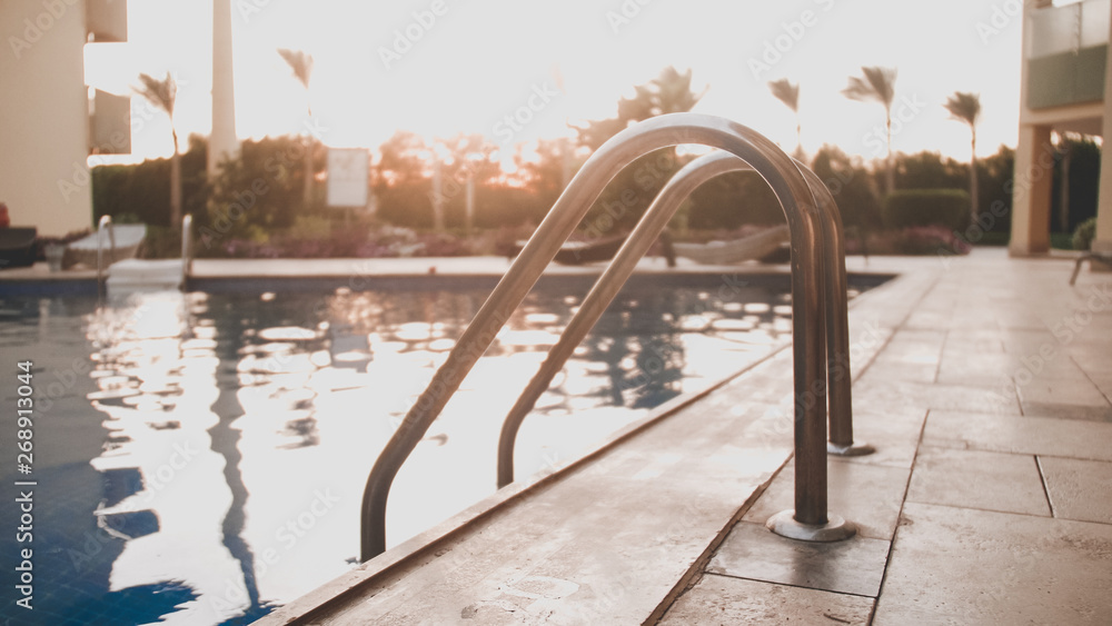 Closeup toned image of metal staircase railings in swimming pool against amazing sunset sky Closeup toned image of metal staircase railings in swimming pool against amazing sunset sky