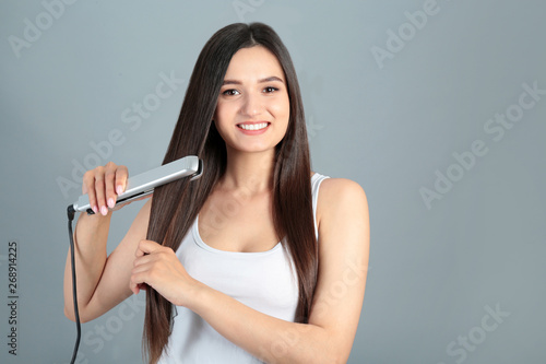 Young woman using hair iron on grey background, space for text