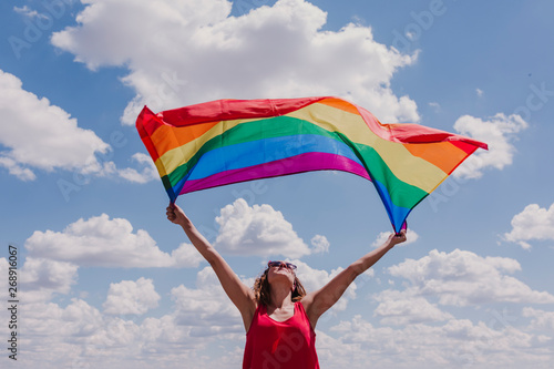 Woman holding the Gay Rainbow Flag over blue and cloudy sky outdoors. Happiness, freedom and love concept for same sex couples. LIfestyle outdoors