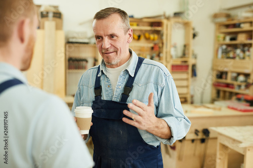 Portrait of senior carpenter talking to worker during coffee break in workshop, copy space
