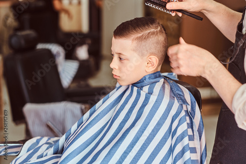 Funny little boy is preparing for the school at trendy barbershop with mature hairdresser.