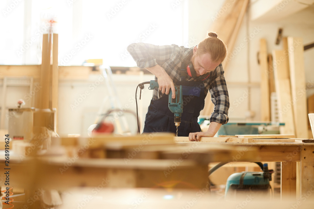 Waist up portrait of red haired carpenter drilling wood while working in joinery lit by sunlight, copy space Waist up portrait of red haired carpenter drilling wood while working in joinery lit by sunlight, copy space