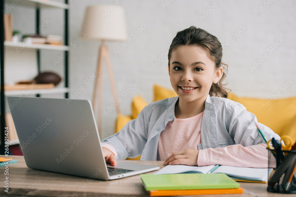 happy child looking at camera while sitting at desk and using laptop at home happy child looking at camera while sitting at desk and using laptop at home