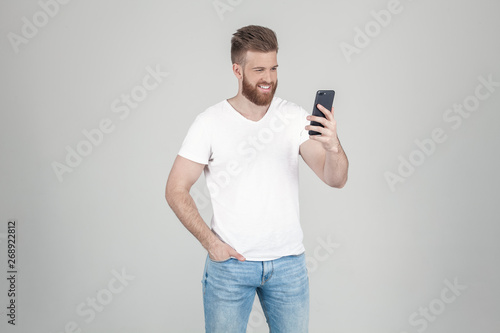 A satisfied man with a lush ginger beard. Smiling to the phone. dressed in casual clothes. stand in front of a white background