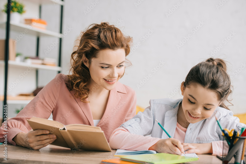 beautiful woman holding book and adorable daughter writing in notebook while doing homework together beautiful woman holding book and adorable daughter writing in notebook while doing homework together