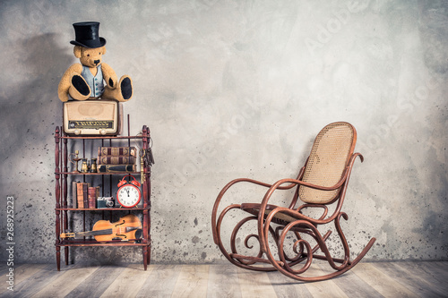 Teddy Bear in cylinder hat on vintage radio, antique books, clock, camera, binoculars, fiddle, keys on shelf, aged rocking chair front concrete wall background. Retro old style filtered photo 