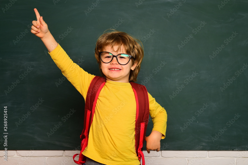 Elementary school and education. Cute boy with happy face expression near desk with school supplies. Funny little boy pointing up on blackboard.