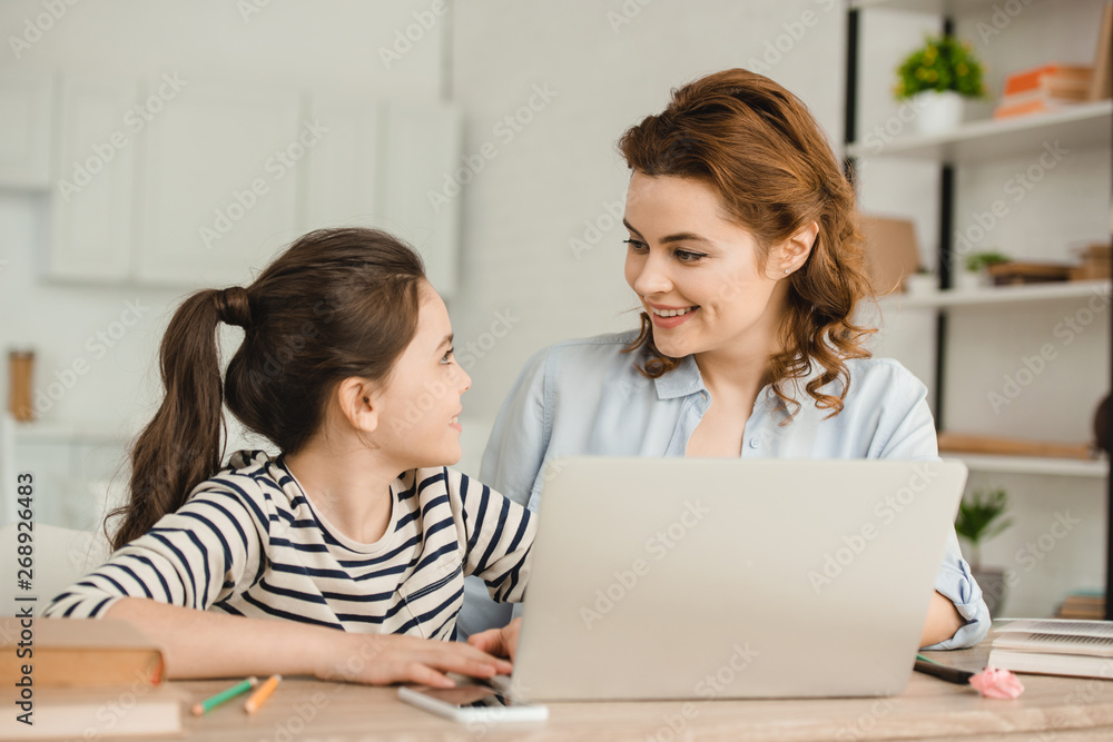 happy mother looking at adorable daughter while using laptop together happy mother looking at adorable daughter while using laptop together