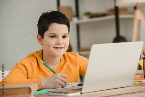 smiling boy writing with pencil and looking at camera while doing schoolwork at home