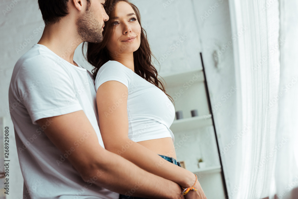 low angle view of man hugging smiling woman in white t-shirt at home low angle view of man hugging smiling woman in white t-shirt at home