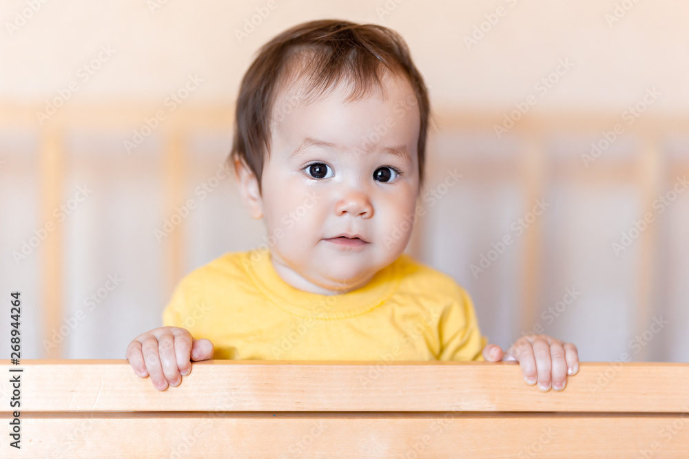 Cute little baby girl with dark hair standing in crib at home in yellow t-shirt Cute little baby girl with dark hair standing in crib at home in yellow t-shirt