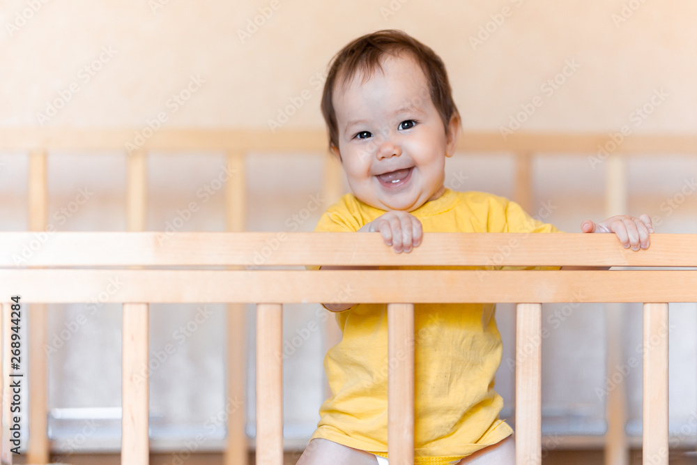 Happy baby girl with dark hair laughing and smiling in yellow t-shirt mixed race kazakh and german standing in cot crib Happy baby girl with dark hair laughing and smiling in yellow t-shirt mixed race kazakh and german standing in cot crib