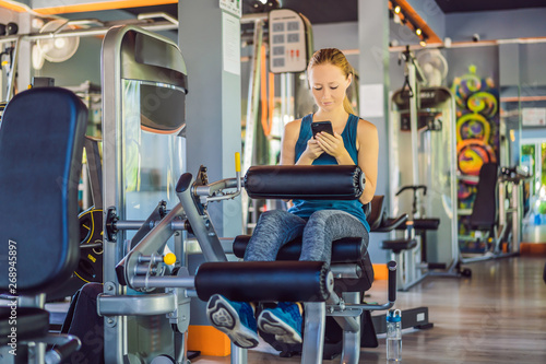 Young woman using phone while training at the gym. Woman sitting on exercising machine holding mobile phone