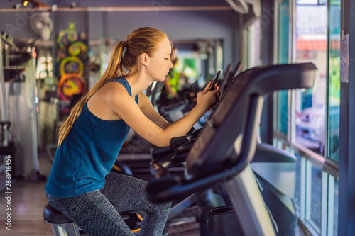 Young woman using phone while training at the gym. Woman sitting on exercising machine holding mobile phone