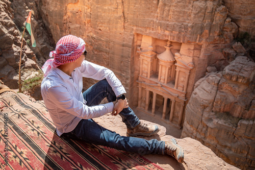 Asian man traveler sitting on carpet viewpoint in Petra ancient city looking at the Treasury or Al-khazneh, famous travel destination of Jordan and one of seven wonders. UNESCO World Heritage site.