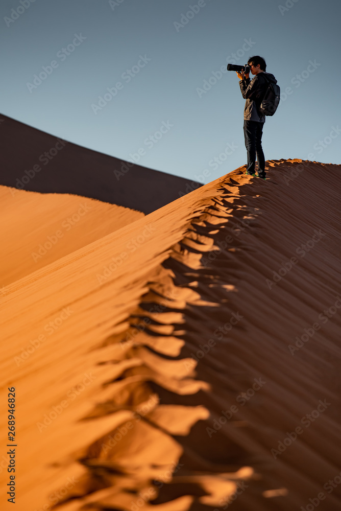 Φωτογραφία Young Asian man traveler and photographer standing on the top of sand dune photographing sunrise or sunset in desert of Namibia, Africa