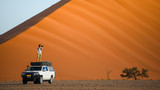 Young Asian man traveler and photographer standing on camper car near orange sand dune. Desert road trip travel concept