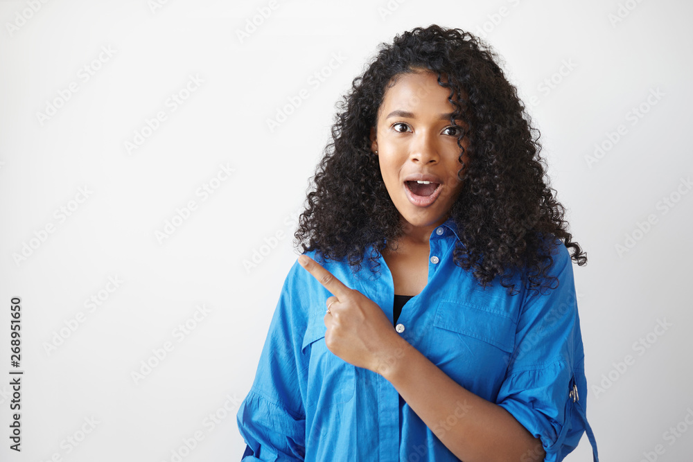Check this out. Waist up shot of beautiful amazed young dark skinned female in blue shirt staring at camera in full disbelief, jaw dropped, pointing fore finger against white copyspace wall Check this out. Waist up shot of beautiful amazed young dark skinned female in blue shirt staring at camera in full disbelief, jaw dropped, pointing fore finger against white copyspace wall