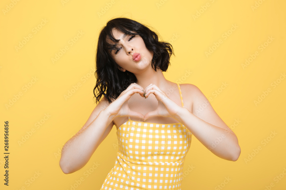 Φωτογραφία Beautiful stylish caucasian female showing heart symbol while kissing and looking at camera dressed in yellow dress isolated over yellow background