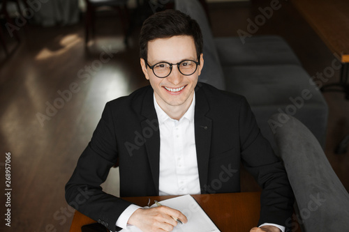 Portrait of a stylish young elegant freelancer looking at camera smiling while sitting at a desk taking notes in his notebook. photo