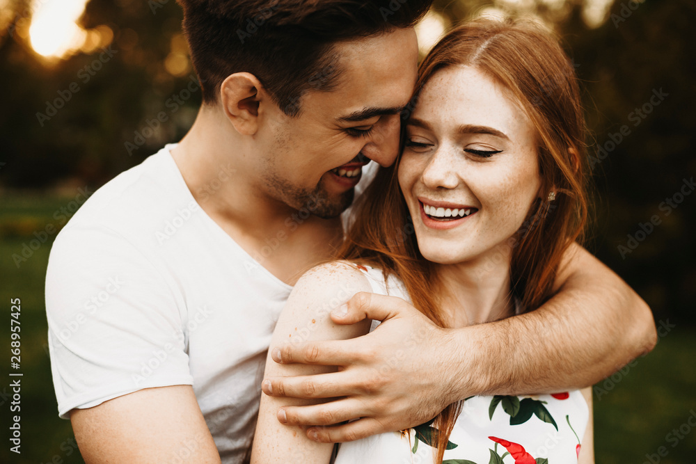 Portrait of a amazing couple laughing while man is embracing from back her girlfriend with red hair against sunset while dating in their vacation time. - obrazy, fototapety, plakaty 