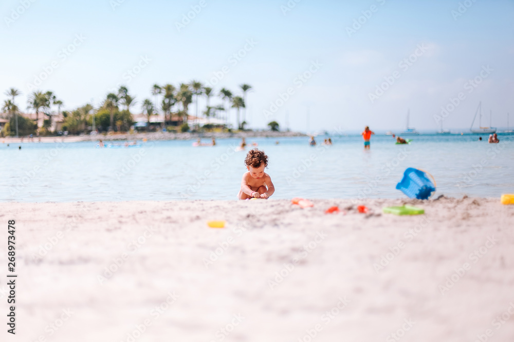 Cute little curly baby play with water and sand on beach at the seaside. Enjoying a lovely summer day. Vacation concept