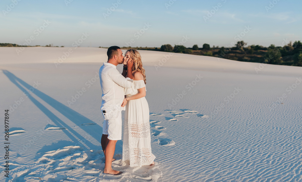 Young beautiful couple of blonde and tanned brunet guy are standing at sunset and kissing. Romantic lovers walk on the sand in white clothes at sunset. Love, honeymoon and idyllic relationships. Young beautiful couple of blonde and tanned brunet guy are standing at sunset and kissing. Romantic lovers walk on the sand in white clothes at sunset. Love, honeymoon and idyllic relationships.