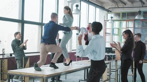 Colleagues fun dancing and jumping on the table, singing in a megaphone and scatter money. Managers celebrate success. Corporate party business team. Modern trendy office interior. Office life