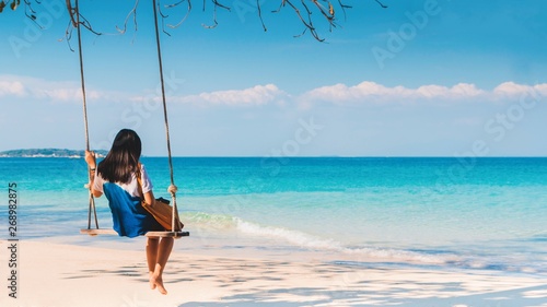 Happy Asian woman sitting on a swing on the white sand beach by the blue sea background in summer of Thailand.Travel in holidays concept.