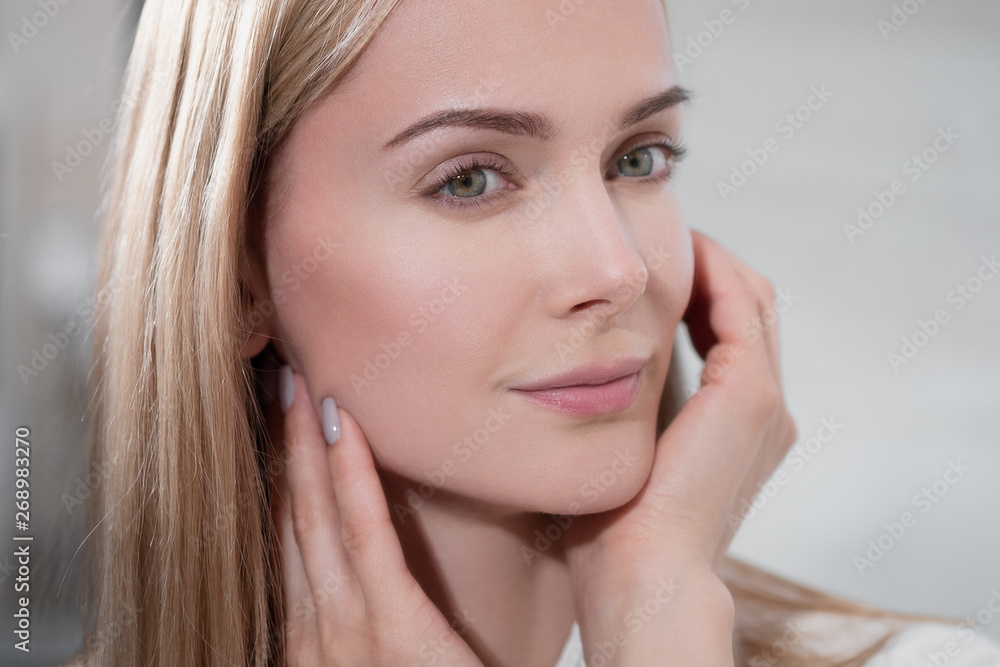 Attractive middle-aged blond woman in a white bathrobe stands in the bathroom by the mirror. She touches skin and smiles. Attractive middle-aged blond woman in a white bathrobe stands in the bathroom by the mirror. She touches skin and smiles.