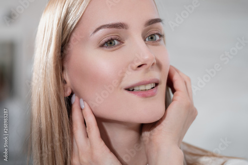 Attractive middle-aged woman a blonde in a white coat stands in the bathroom by the mirror. She touches skin and smiles. Beautiful white teeth lips and eyes.