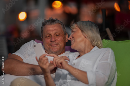 Happy mature couple spending evening together outdoor. They relaxing in chaise longues and looking at photos on mobile