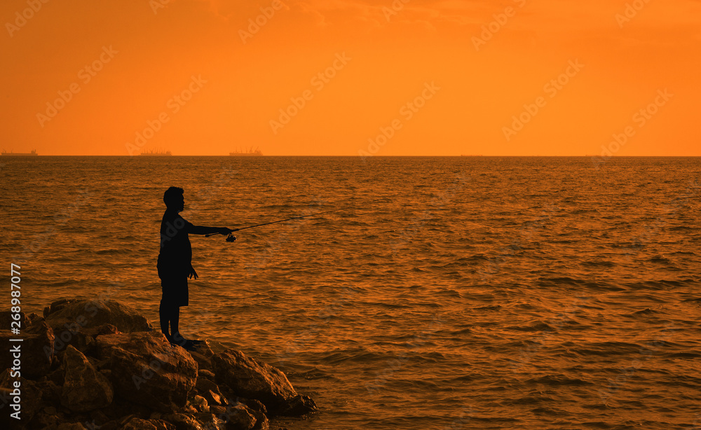 Fisher man fishing with spinning rod on a sea . Young men Fishing in the reservoir At sunset . Silhouette of a fisherman at sunset. Fisher man fishing with spinning rod on a sea . Young men Fishing in the reservoir At sunset . Silhouette of a fisherman at sunset.