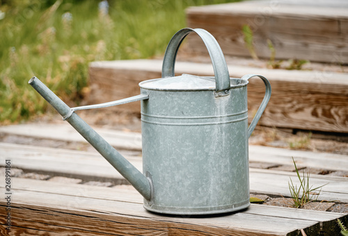Old metal watering can standing on the wooden stairs of a terrace in the garden with grass and plants in the background photo
