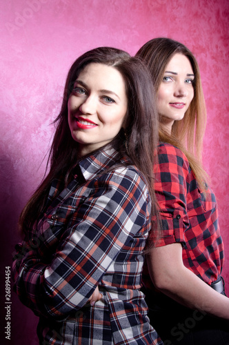 two young beautiful women in plaid shirts stand with their backs to each other on a pink background