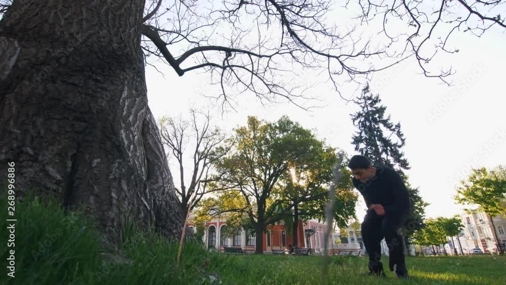 Young man parkour athlete doing backflip jump from the tree in city park, slow motion, low angle view Young man parkour athlete doing backflip jump from the tree in city park, slow motion, low angle view